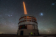Central in the image is the Unit Telescope 4 of ESO’s Very Large Telescope, standing in front of a starry night sky. The telescope is cylindrical in shape, with several openings on its side. From the top of the structure, four orange laser beams shoot out into the sky, converging at a single point. The sky is full of stars. Some of the stars are much brighter than others and there is a particularly bright area off to the right of the telescope. Most of the stars are white or blue in colour, although some have a more orange colour, particularly those near the horizon.