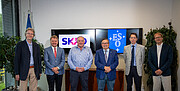 This image shows ESO and SKAO representatives at the signing ceremony standing and looking at the camera while smiling. They are smartly dressed, most wearing suit jackets and ties. The image was taken in an office setting. Behind the line of gentlemen, two TV monitors are displaying the SKAO (left) and ESO (right) logos. There are also two plants on either side of the office, behind the men.