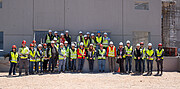 Around 30 people are standing in two lines in front of a large concrete wall, wearing high-visibility jackets and hard hats.