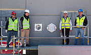 Four people in high-visibility jackets and hard hats are standing on scaffolding in front of a large grey concrete wall. The metallic, square time capsule and accompanying hexagonal plaque are fitted into the wall between the group.