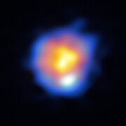 A bright, colourful blob sits at the centre of the image, in stark contrast to the dark background. From the centre outwards, there are amorphous layers of yellow, pink and blue. A white spot stands out at the bottom of the pink ring. Wispy tendrils of blue fade out into the background.
