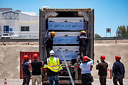 Looking in the open back door of a lorry, or truck, three large white boxes fill the lorry bed, surrounded by white air bags. Two people with protective helmets stand in the truck bed, while six others stand on the ground below, everyone with their backs to the camera. In the distance, brown ground juts up with a white building sitting atop under a blue sky.