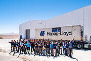 A group of about 40 people stand on beige concrete in front of a large white building and a lorry, or truck, labelled Hapag-Lloyd. There are many hands in the air and many expressive faces. This scene is complemented by a light but vivid blue daytime sky.