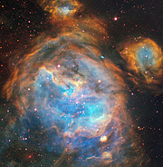 A mesmerising image of wispy orange clouds surrounding a bright blue-purple region. The clouds are rounded in shape and give the impression of cosmic bubbles floating through space. The dark background of the image peppered with the bright spots of stars, most with a slight red tinge to their hue.