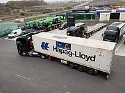 On dark grey concrete, a white lorry, or truck, with the words Hapag-Lloyd on the side is parked, looking ready to drive off to the left side of the image. Just behind it, other cars and trailers sit in parking bays. Spanning off into the distance are shipping containers of different colours and brown land.