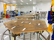 Several hexagonal brown segments are lined up on metal supports in a workshop setting. The upper surface of each is polished and smooth. A few workers can be seen in the background in blue lab coats and masks.