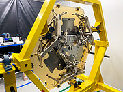 In a workshop setting, a large yellow hexagonal metal frame is positioned vertically with gold and silver mechanical structures suspended inside it.