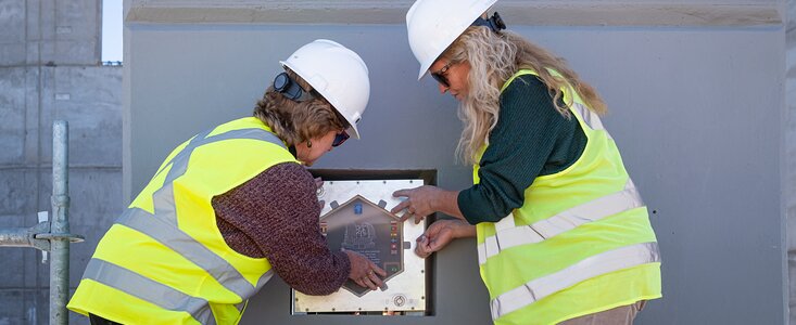 Two people in high-visibility jackets and hard hats are standing on either side of a shiny, metallic square box – the time capsule – placing it into a grey concrete wall.
