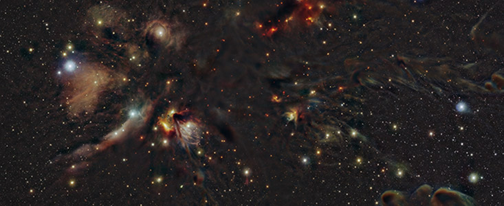 This image shows stars and clouds of gas and dust distributed over a dark background. A prominent cloud of gas and dust can be seen in the upper left part. It features amorphous clouds in red, green, blue and yellow colours. A similar, but smaller, cloud can be seen both in the upper right and lower right part of the image.