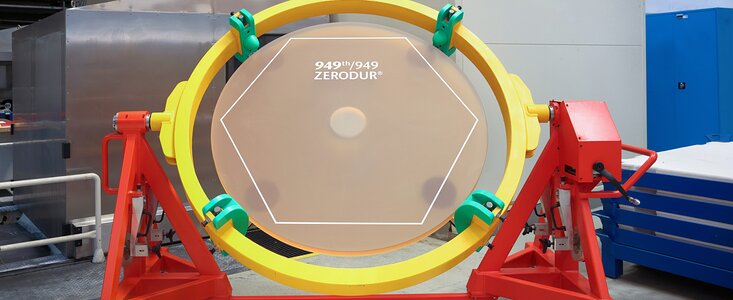 A brown, translucent circle is held up by a yellow and red metallic structure in a workshop environment. A white line traces the outline of a hexagon on the material, and white text reads “949th/949 ZERODUR©”. The name “SCHOTT” in blue letters can be seen on a machine in the background.