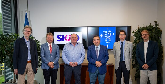 This image shows ESO and SKAO representatives at the signing ceremony standing and looking at the camera while smiling. They are smartly dressed, most wearing suit jackets and ties. The image was taken in an office setting. Behind the line of gentlemen, two TV monitors are displaying the SKAO (left) and ESO (right) logos. There are also two plants on either side of the office, behind the men.