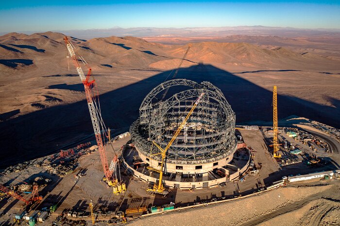 In the foreground, at the centre of the picture, is the Extremely Large Telescope, with its concrete round base and its steel structures of the dome. Around the telescope are trucks, cranes and several construction vehicles. In the background, a large triangular shadow is cast upon the ground by a mountain mostly out of the frame, the mountain atop which the telescope is being built. Further on are light brown mountains, while a light blue sky encloses the top of the frame.