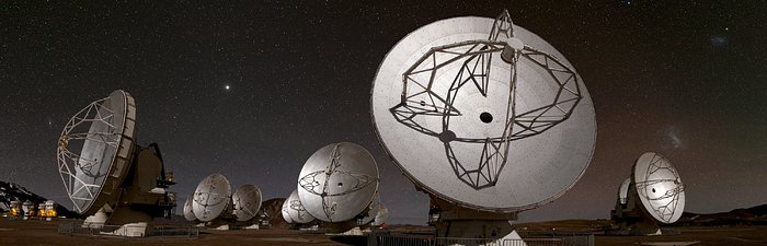 This image shows close to a dozen of the ALMA telescope dishes, all pointing in different directions. The dishes are large, almost bowl-like and and silvery in colou. The image is captured during the night, with a dark sky in the background, and the moon illuminating the dishes.