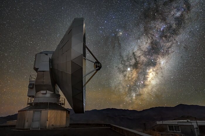 Night image of the Swedish-ESO Telescope with an overall grey and dark blue colour. The telescope is a big antenna seen sideways, taking up most of the left part of the image. To the right of the telescope the Milky Way dominates the sky and contains dark and bright areas. The Chilean mountains shape the horizon above the ground in the lower part of the image.