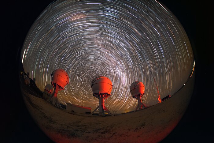 The image is taken through a circular fisheye lens during the night and shows the three domed telescopes positioned at the left, centre and middle-right of the image. Each telescope is illuminated from the right hand side by an orange light. Behind the telescopes, in the upper three quarters of the image, are many curved streaks of white, blue and yellow star light on the dark background of the night sky. The curves of light are all centred around a point just to the left of the central telescope.