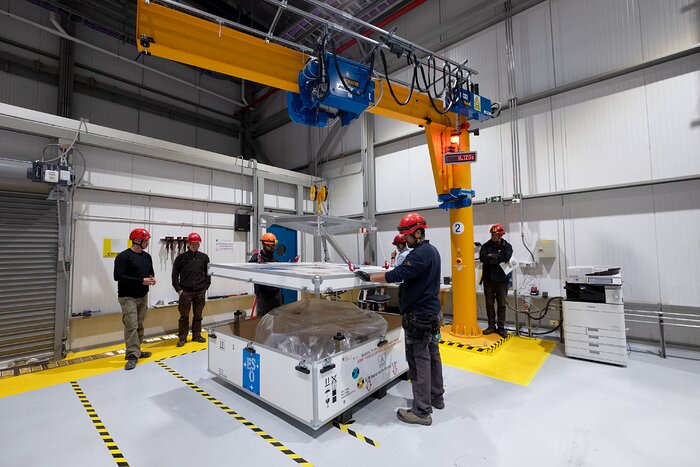 Inside a large warehouse, six people with red protective helmets stand around a large white box under a yellow and blue crane. The arm of the crane is hovering over the box with a pulley lifting the square lid off the box. The floor, mostly grey, has spots of bright yellow paint. The box is labelled with the ESO logo — four stars around the letters E, S, O on a blue background — and inside is a plastic sheet wrapped around a large unknown object.