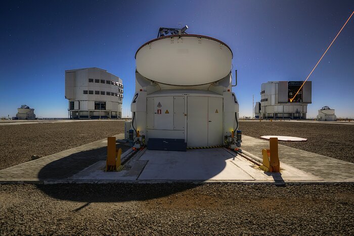 Looking out across a brown, pebbly plateau, five white telescope structures are seen. In the front and centre, a rounded dish with a bulky base sits upon a brown-grey concrete slab, a large shadow protruding from it. A geometric telescope dome stands on each side towards the background, large in relative size but small in this perspective. Dishes matching the central one sit further out, with one at each edge of the photo. Above the domes is a vivid blue sky, glowing lighter near the ground as if illuminated from the telescopes.