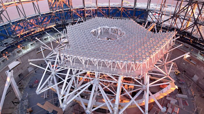 The image shows a partially-constructed white hexagonal lattice structure with a honeycomb-like design, in the interior of a metallic dome structure with some blue cladding on the exterior. In the centre of the lattice structure, there is a hexagonal hole in the structure. The Chilean desert at sunset can be seen in the landscape outside of the metallic dome structure.